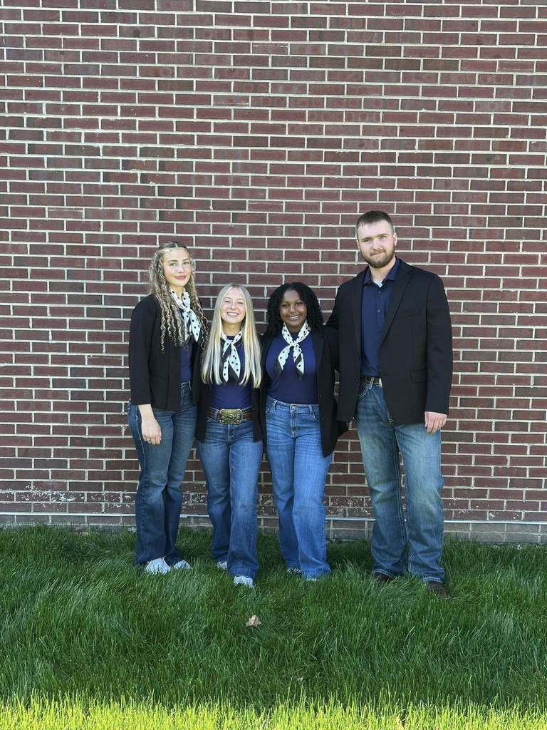 Four individuals stand on grass, wearing casual clothes. They are in front of a brick wall.