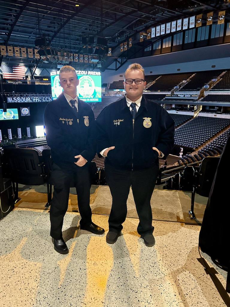 Two men in matching uniforms and ties pose for a picture in a large, empty stadium.