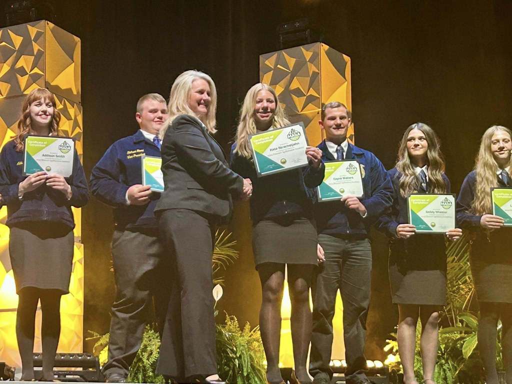 Six individuals on stage, holding certificates, smiling and looking at the audience with a yellow and black background.