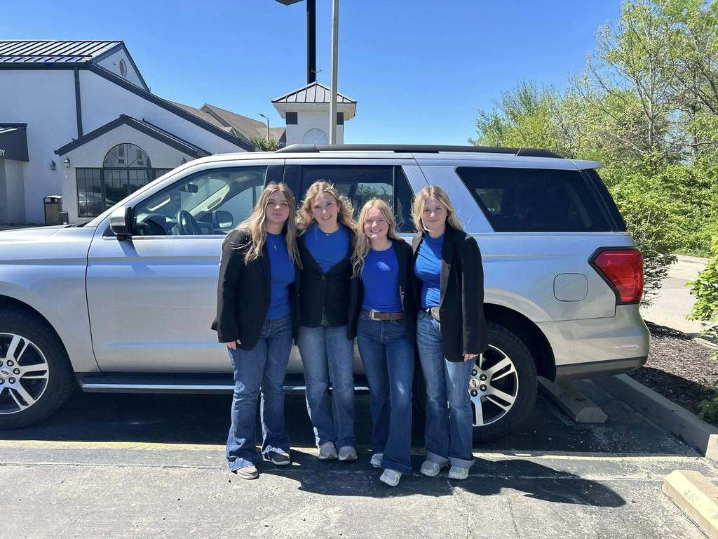 Four women in blue shirts and jeans stand in front of a silver SUV parked in a lot.
