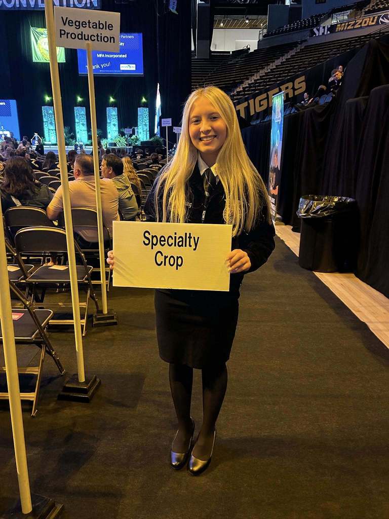 Woman with blond hair holding a sign that says "Specialty Crop" in an auditorium with chairs and banners.