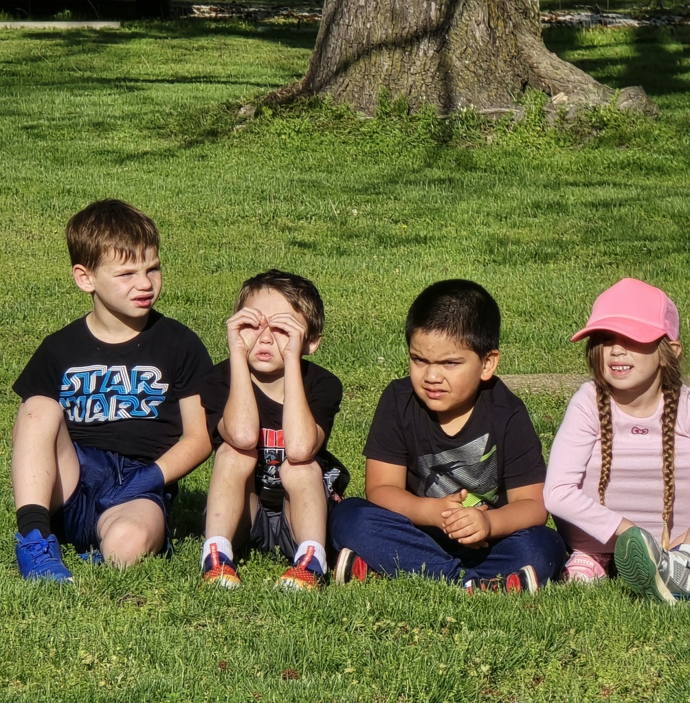 Four children sit on grass near a tree, one in a pink hat. Two boys wear black shirts, one with "Star Wars".