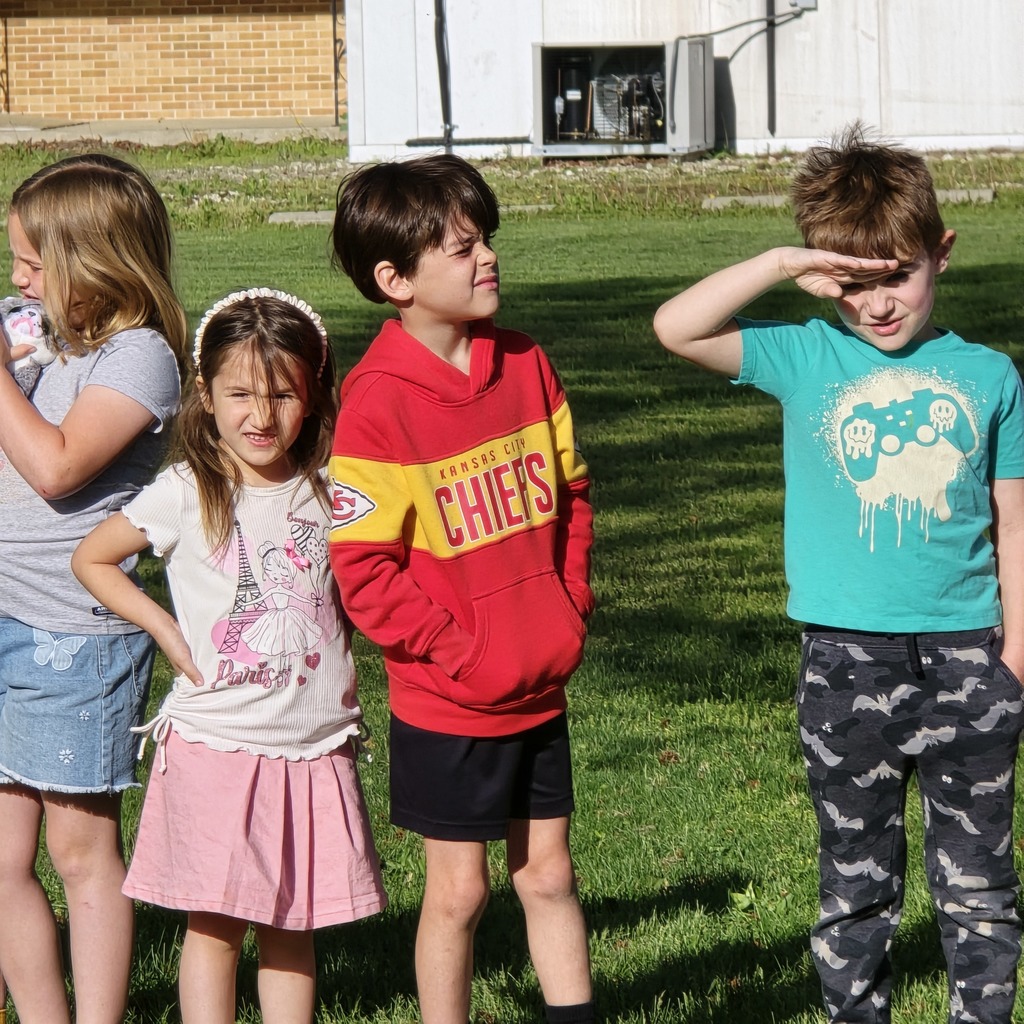 Four children, two girls and two boys, stand on grass. The boy on the right wears a green shirt with a cloud design.