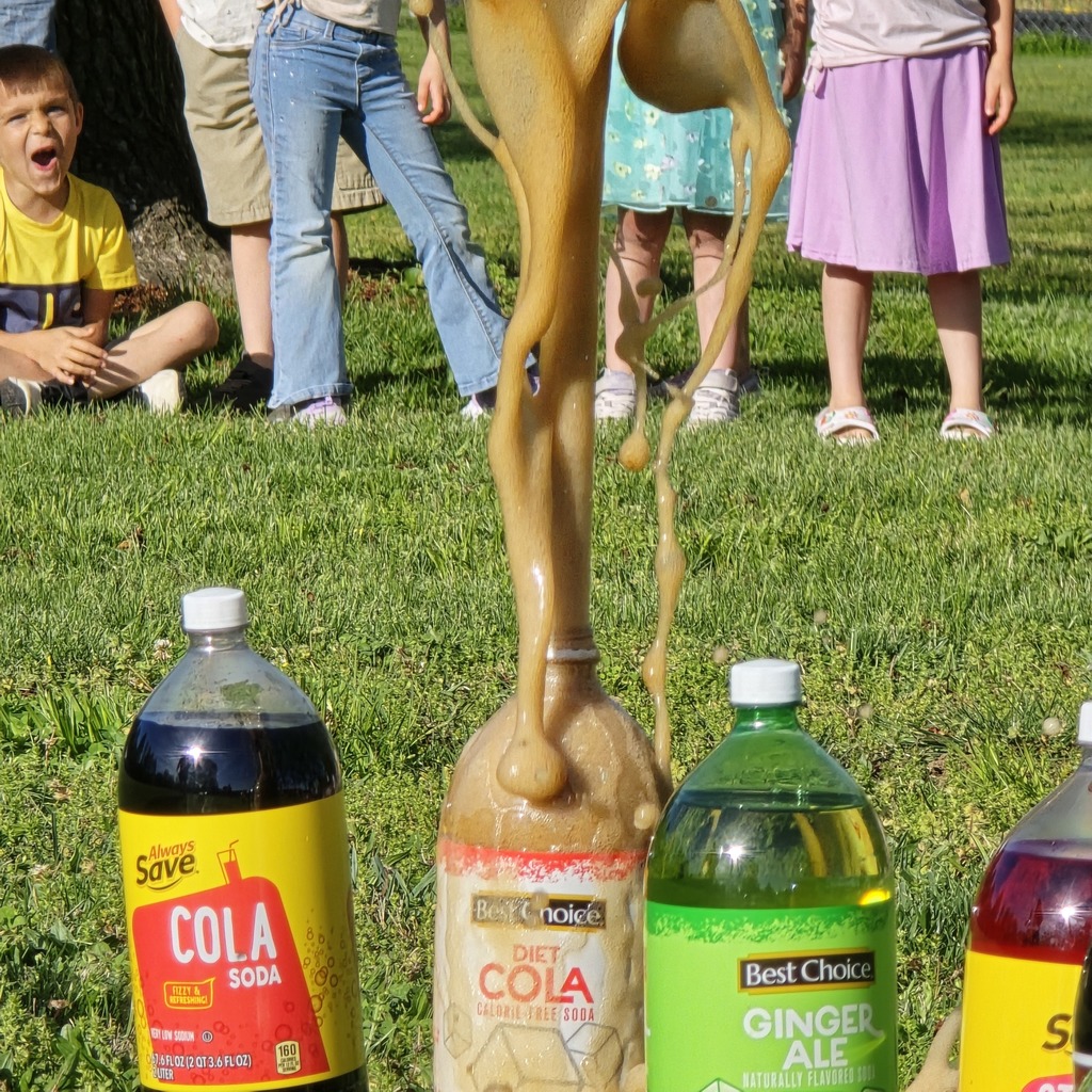Four soda bottles on grass, one spilling, with people in the background.