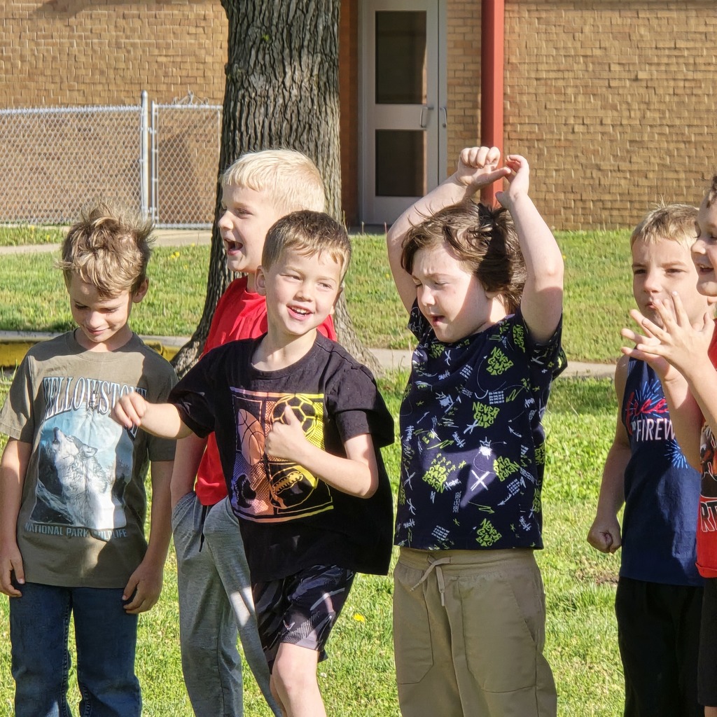 Five children in casual clothes stand on a green lawn. Two have their arms raised. Behind them is a tree and a building.