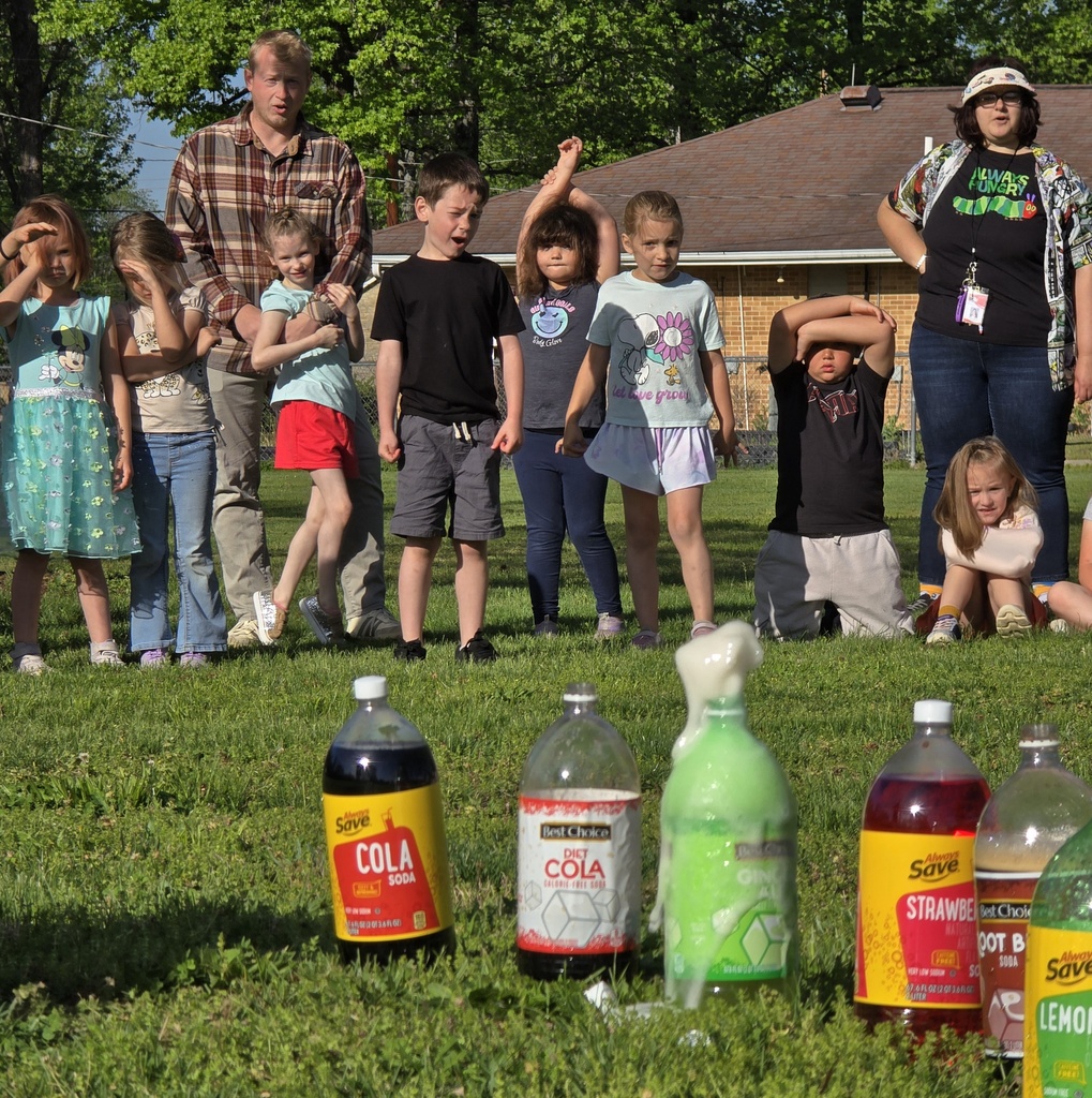 Group of children and adults standing on a lawn with various soda bottles on the ground.
