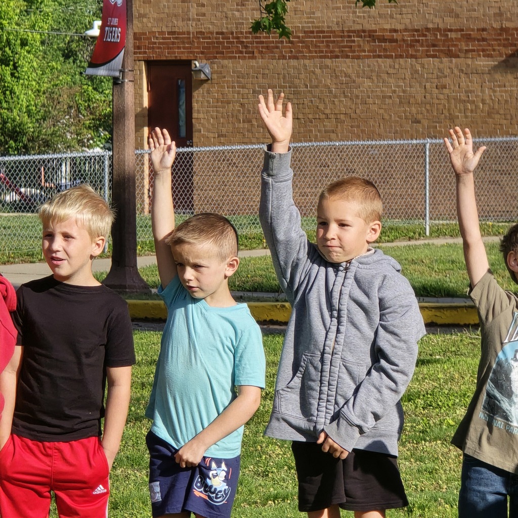 Five young boys in different colored shirts and pants standing in a grassy area in front of a brick building.