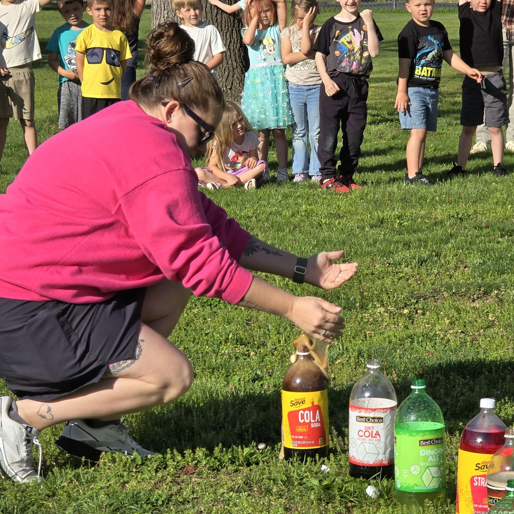 Person in pink shirt crouching on grass, holding a bottle. Children in the background stand and watch.
