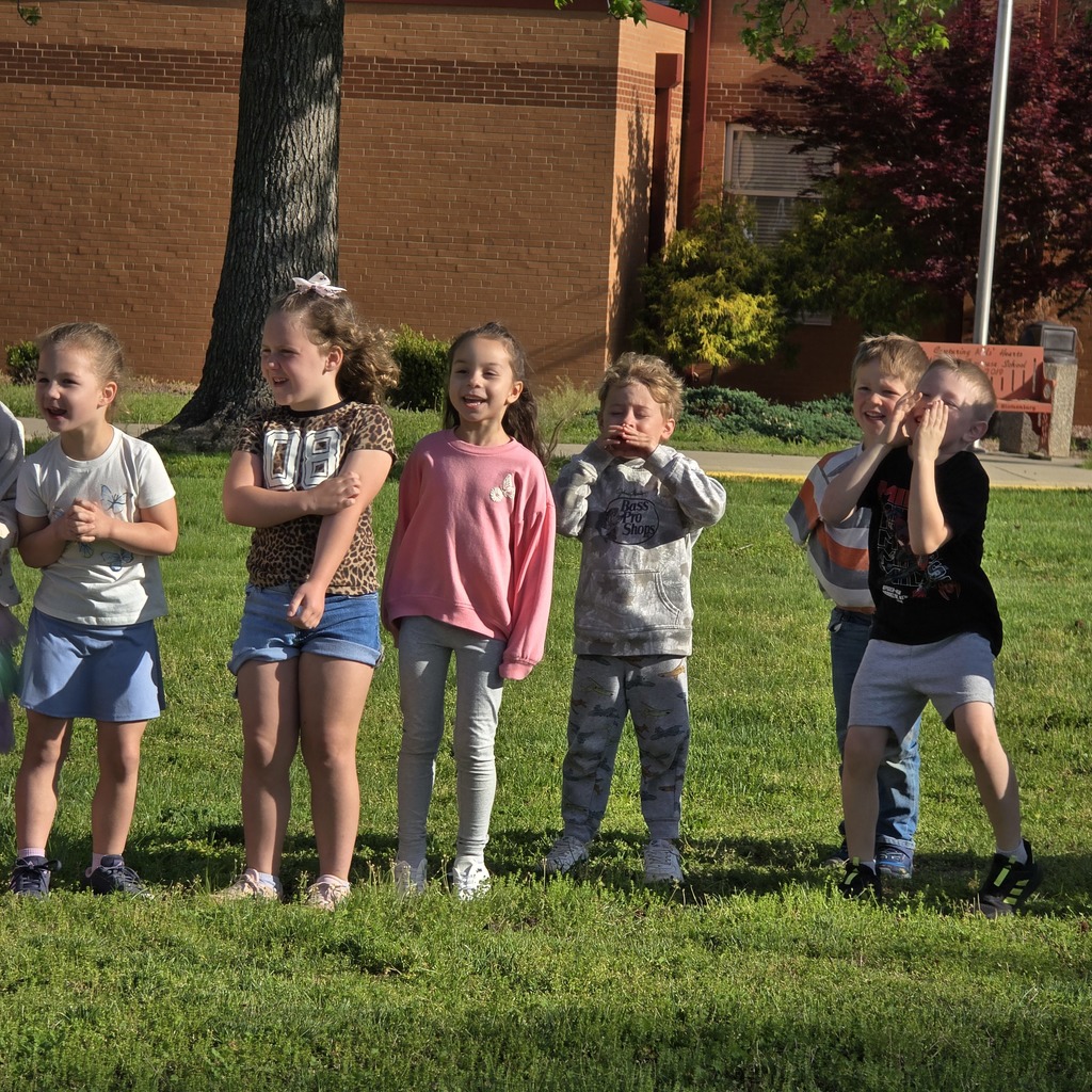 A group of children stands on a grassy area, some looking at something in front. Trees and a building in the background.