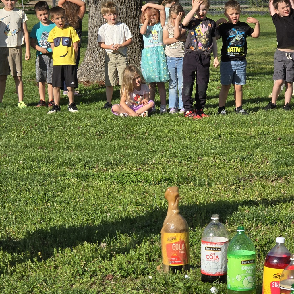 Kids in casual clothing stand and sit on a lawn with a tree. Four bottles are in the foreground.