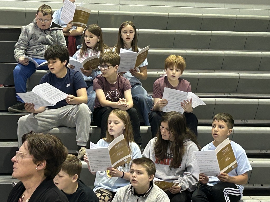 Group of children, some seated on bleachers, holding papers, possibly rehearsing for a performance.