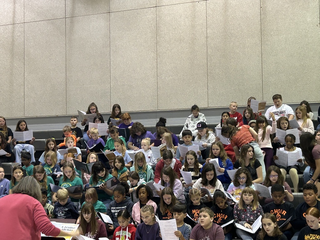A group of students sits in bleachers, holding music sheets. A teacher in pink stands at the front, leading them.