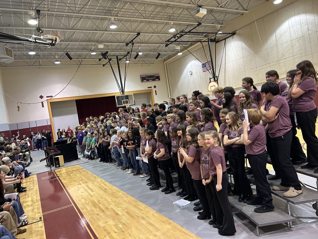 A group of students in purple shirts stands on stage in a school gym with a wooden floor.