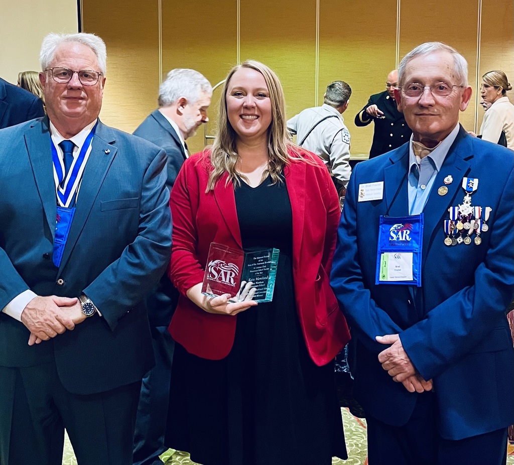 Three individuals, two men and one woman, stand together. The woman holds a trophy, smiling. Both men wear suits and medals.