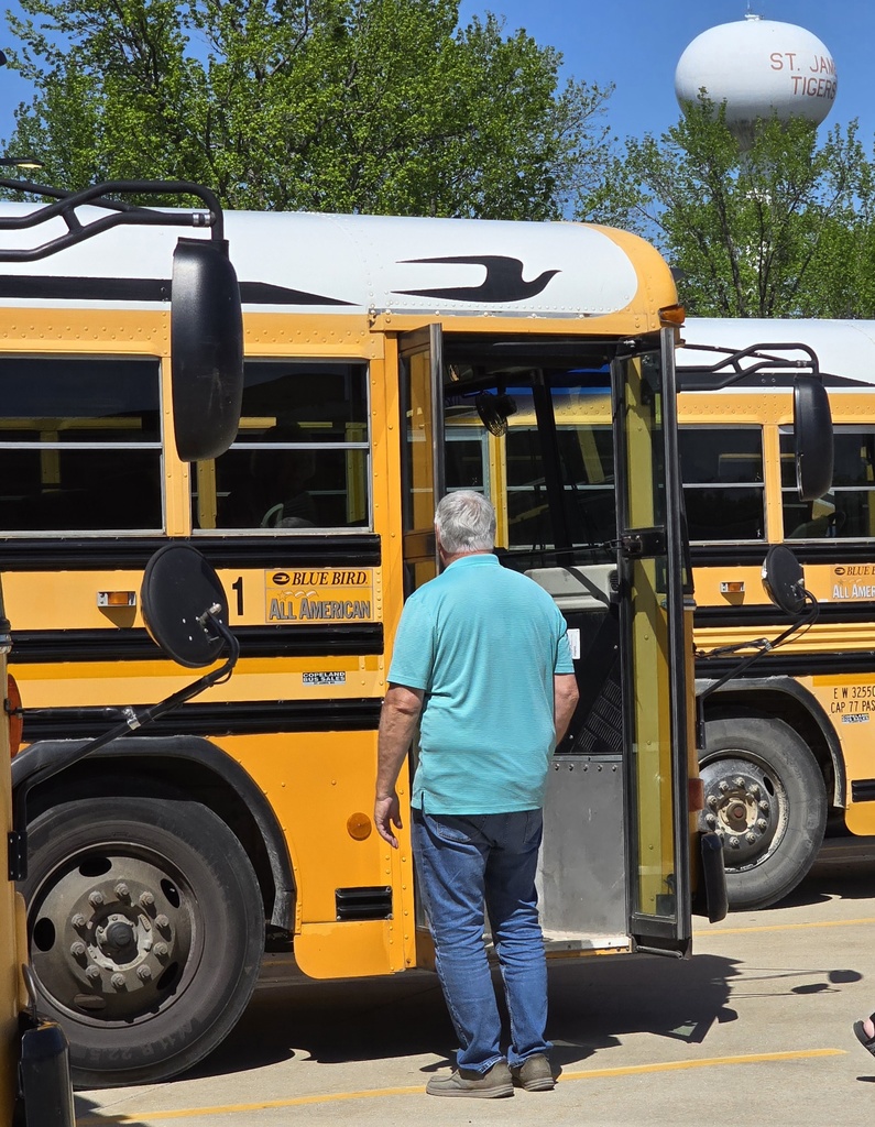 A person boards a school bus in a parking lot with green trees and a water tower in the background.