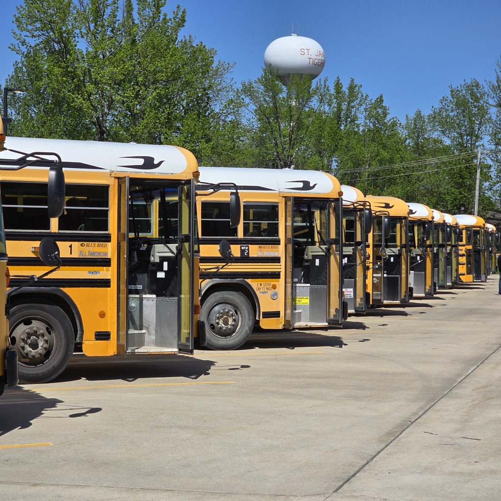 Row of yellow school buses parked in a lot with trees and a water tower in the background.