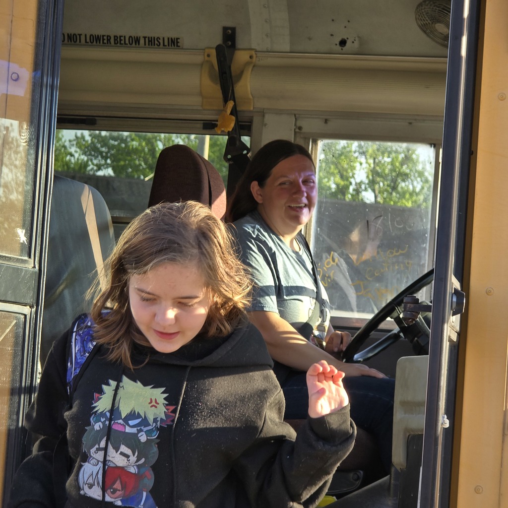Inside a school bus, a woman sits in the driver's seat, smiling. A girl stands in the aisle, smiling and looking forward.