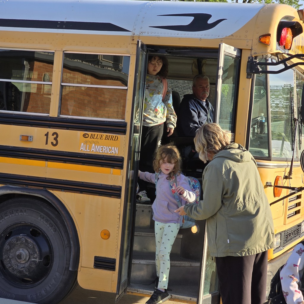 A yellow school bus with an adult and two children boarding. One child wears a pink outfit.