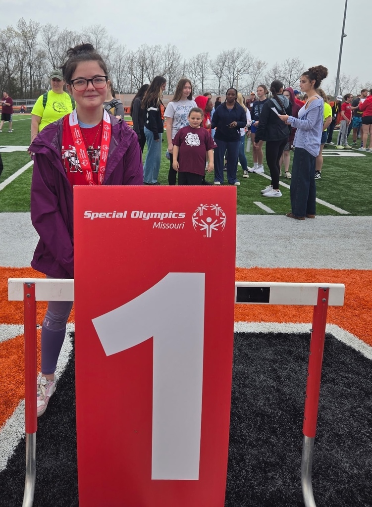 A woman stands in front of a red sign numbered "1" on a field. She wears a medal and glasses.
