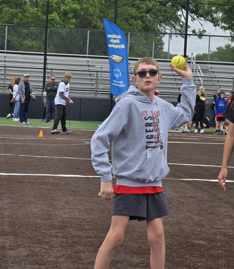 A person in a gray sweatshirt and sunglasses holds a ball, standing on a dirt field. Behind, others stand near bleachers.