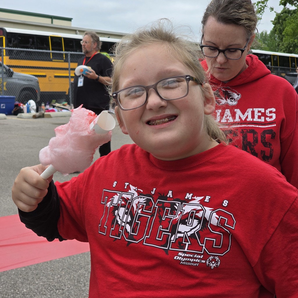 A smiling girl in a red Tigers shirt holds cotton candy. Behind her, a woman in a red hoodie and glasses, a bus, and a pickup truck.