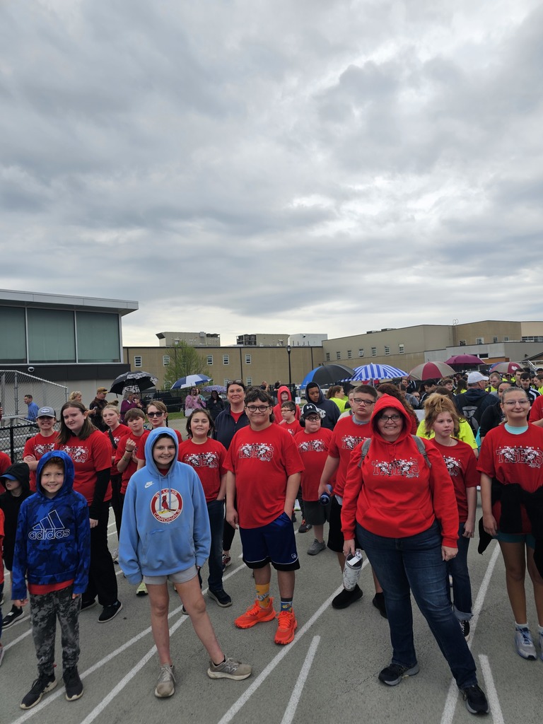 A group of people in red t-shirts, some wearing hoodies and hats, standing on a track, with cloudy skies in the background.