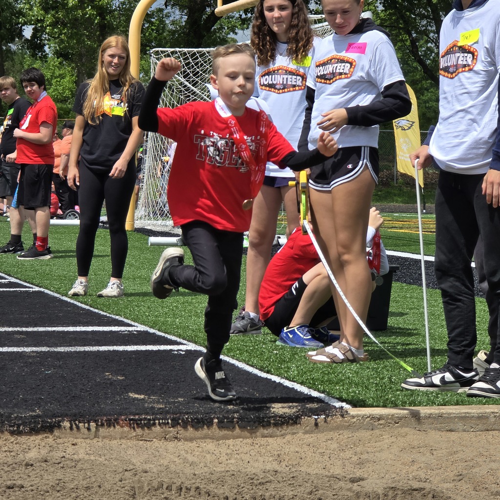 A boy jumps on a track with a blurred background of other people, some wearing white shirts.