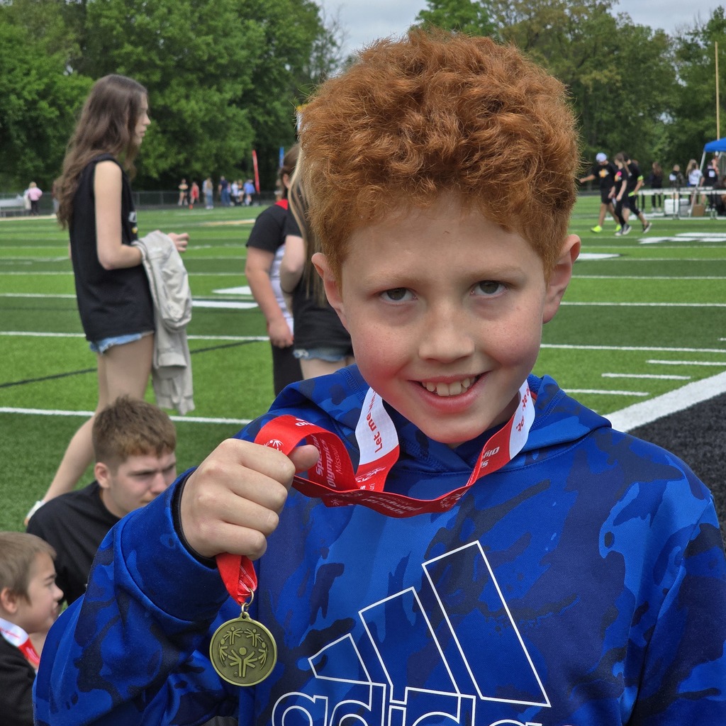 A young boy with red hair holds a medal and smiles at the camera. Behind him, people walk on a football field.