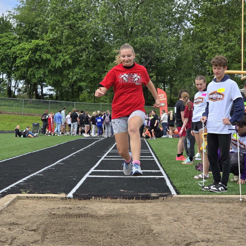 An athlete leaps over a hurdle on a track while spectators watch from behind. Trees are visible in the background.