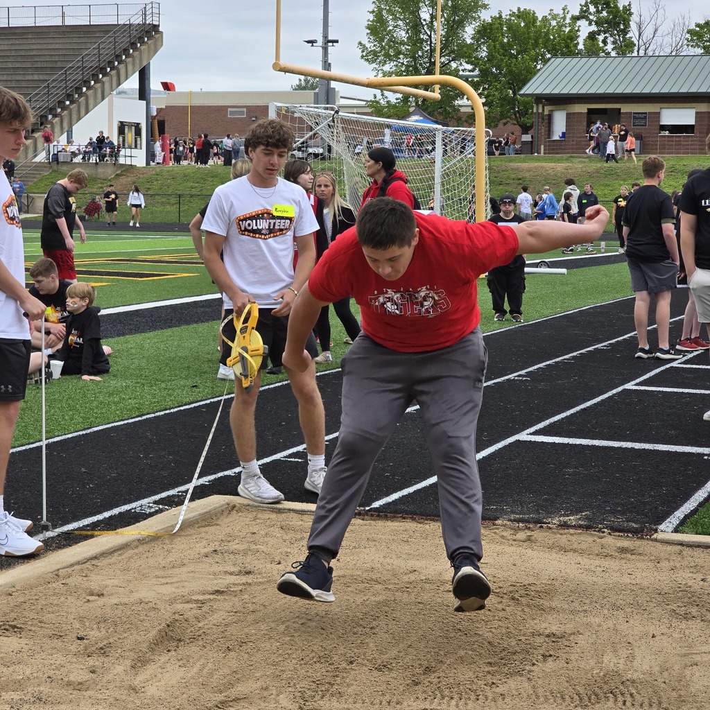 A person in a red shirt and gray pants is jumping on a track. Behind, others are watching.