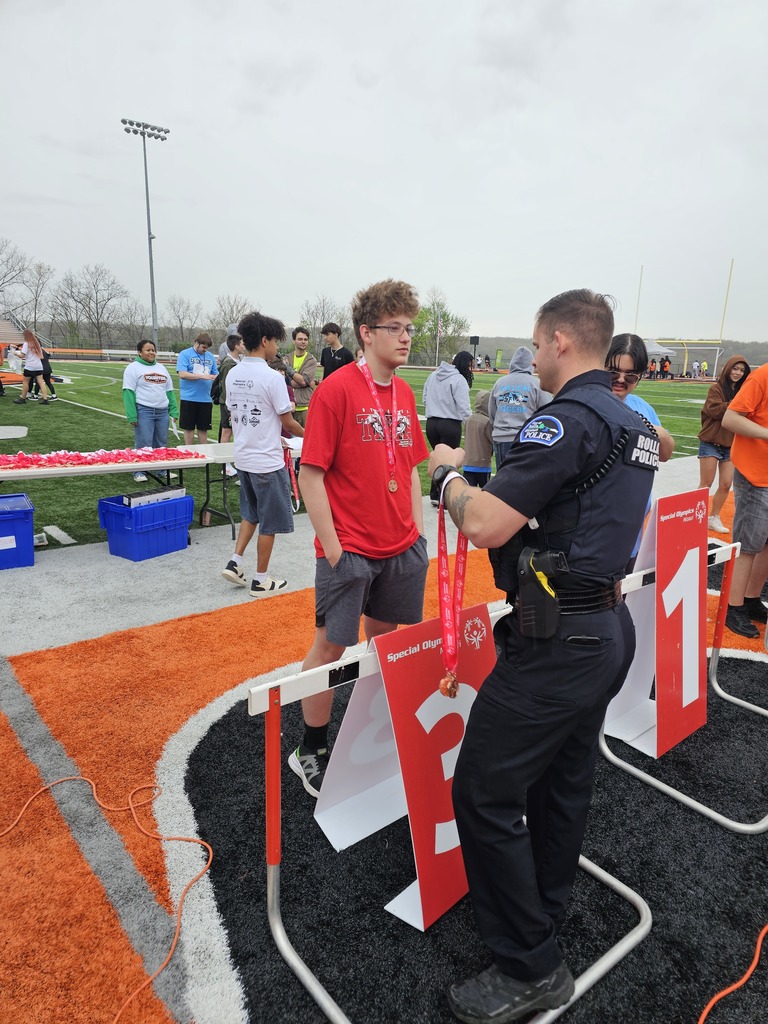 A police officer hands a medal to a boy at an outdoor track. People stand behind them.