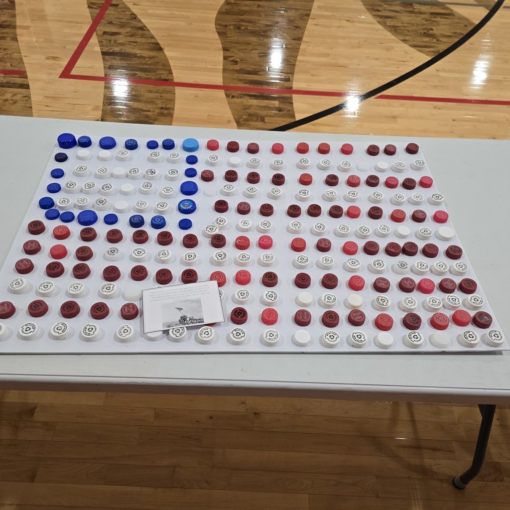 A table displays a flag made from colored bottle caps on a basketball court. Blue, red, and white caps create a design.