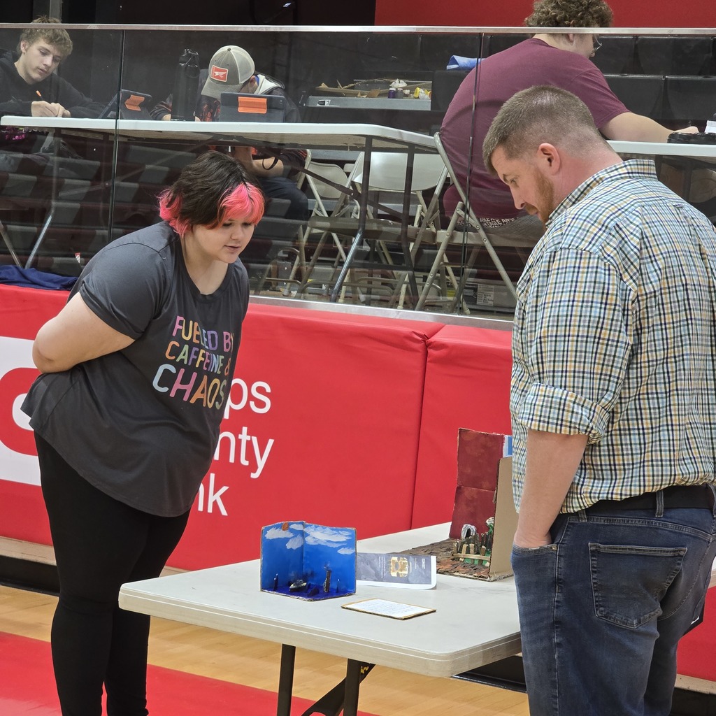 A woman with pink hair stands near a table with a box on it, while a man in a plaid shirt watches.