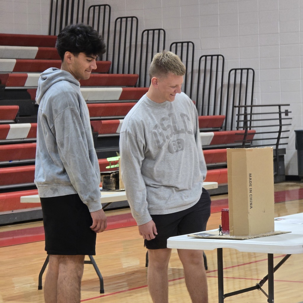 Two men stand by a table with a box. One wears a gray hoodie and black shorts. Behind them, bleachers and a wall.