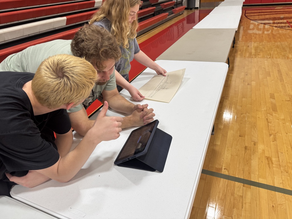 Three people in a gym, looking at a tablet on a table. A woman stands behind them.
