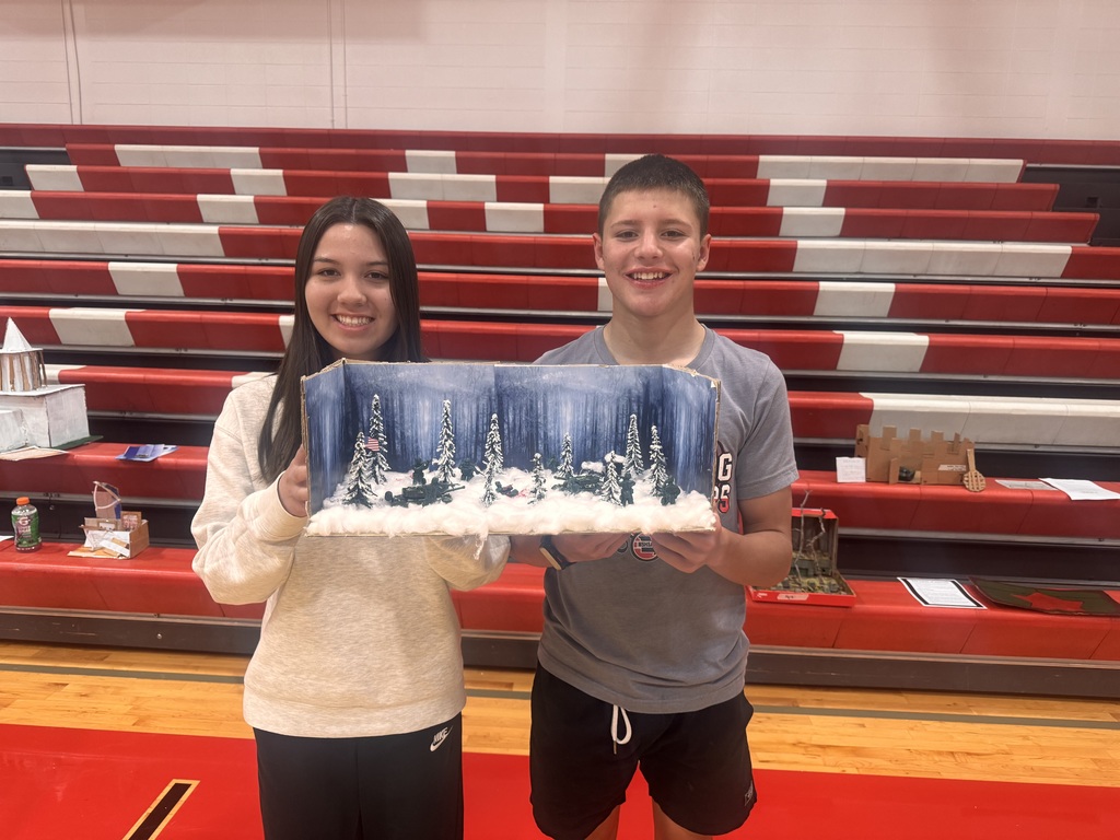 Two individuals in a sports facility, holding a diorama of a snowy landscape with a Christmas tree.