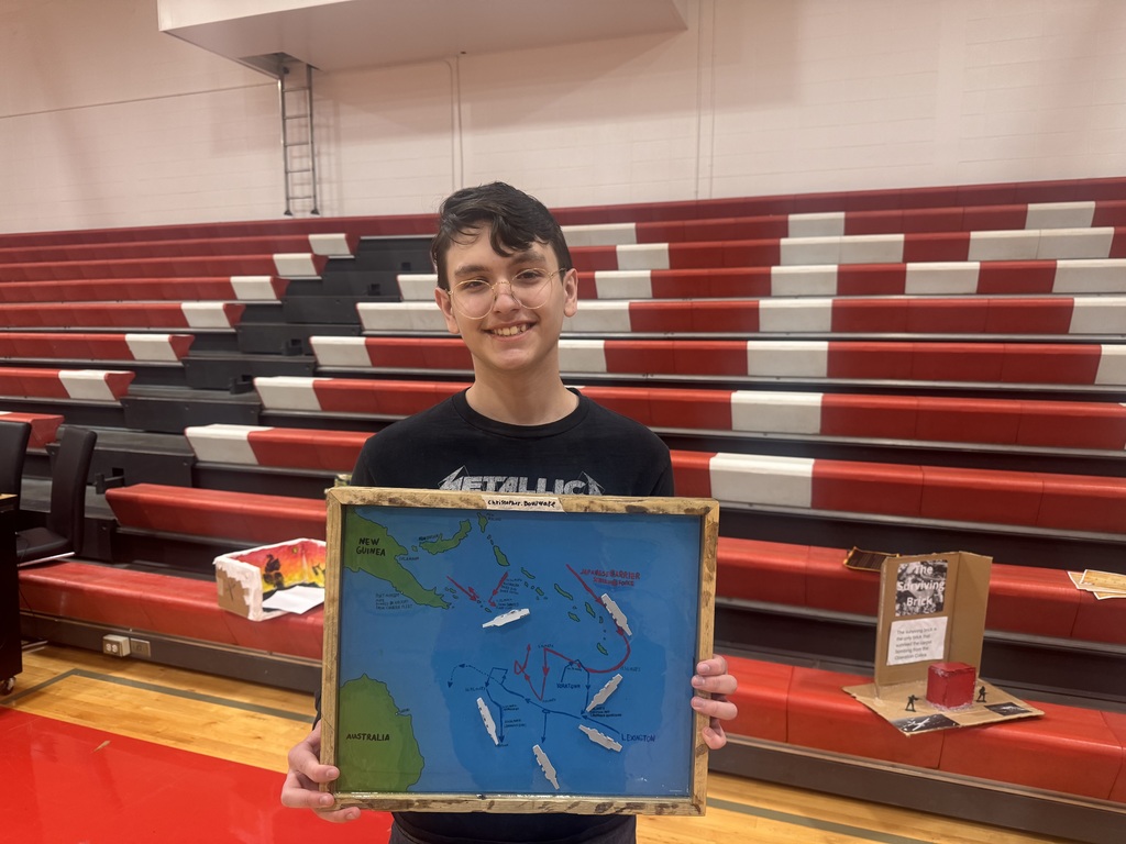 Person standing in a gymnasium holding a framed map with ships depicted. Behind them is a partially empty red and white bleacher.