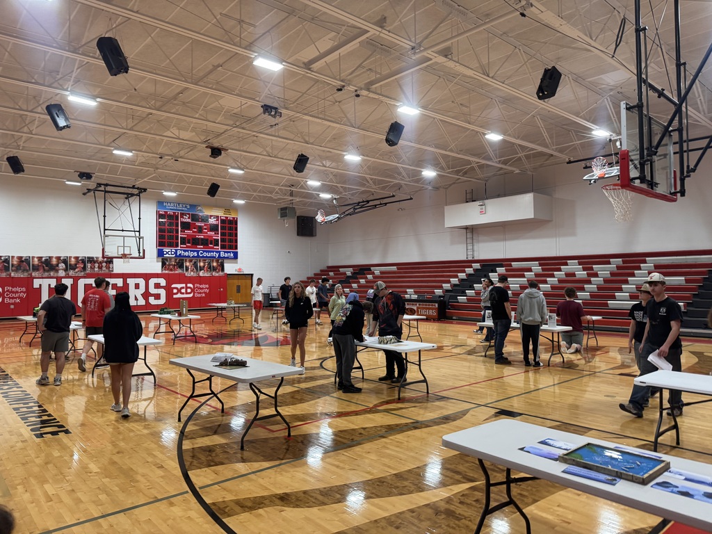 A gymnasium has several tables set up, with a basketball hoop visible in the background. People stand around the tables.