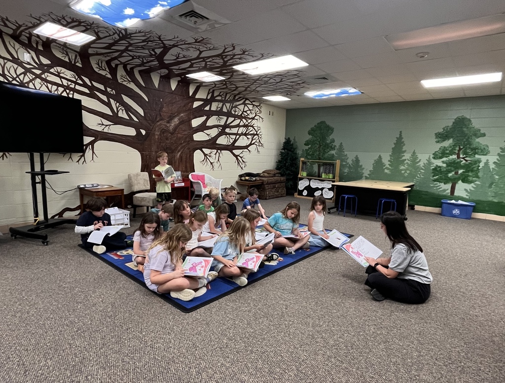 Kids sitting on floor with a teacher, holding books and looking at papers. Background features a tree mural.