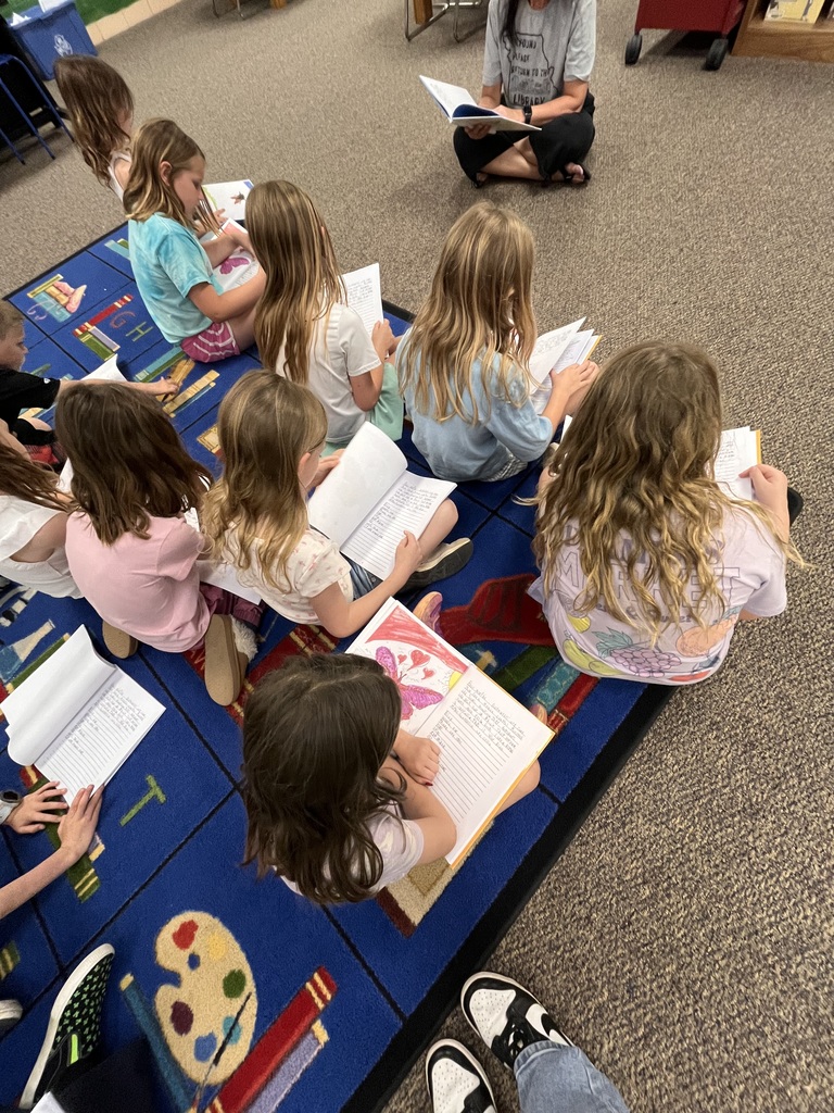 Children sit in a circle on a carpeted floor, reading books. One child holds a book while another holds a paint palette.