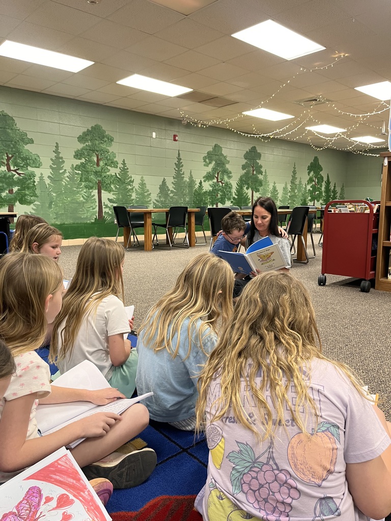 A woman reads a book to young children in a classroom with a tree mural on the wall.