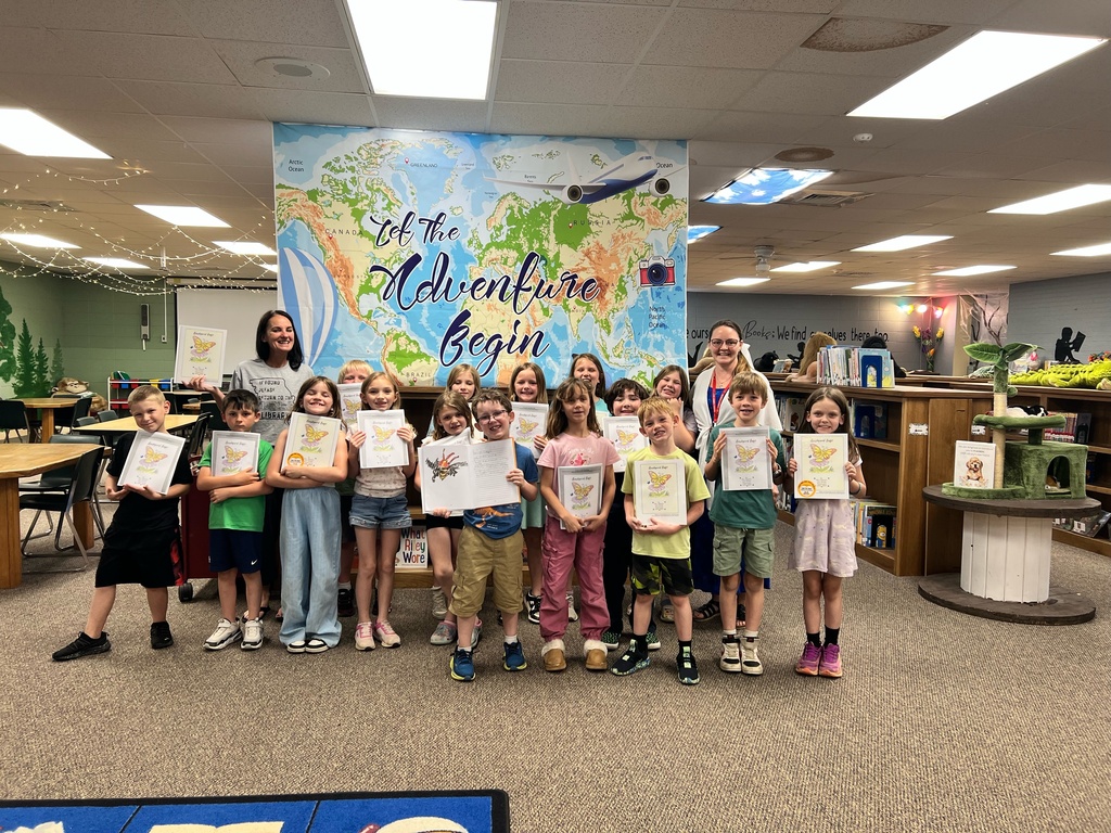 A group of children stands in a classroom, holding certificates. A mural with the words "The Adventure Begins" is behind them.