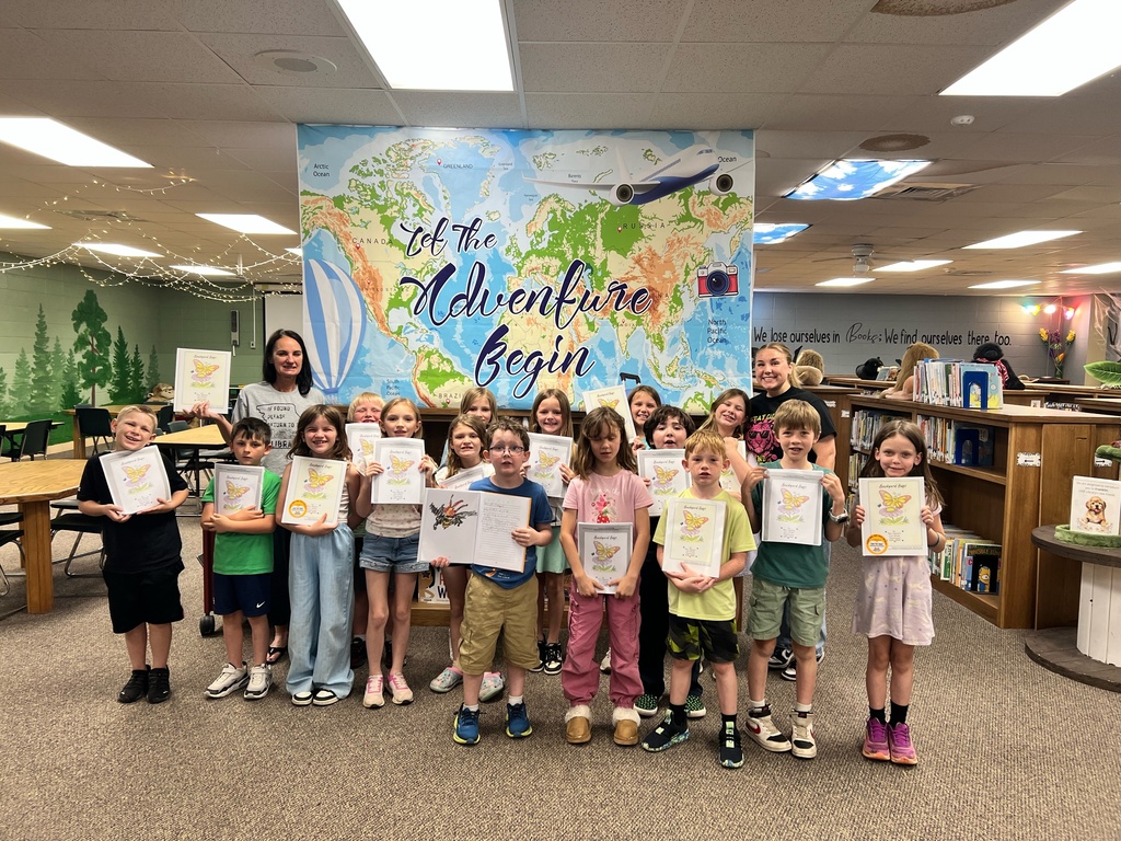 Children stand in a row holding certificates. Behind them is a large world map on the wall and a bookshelf.