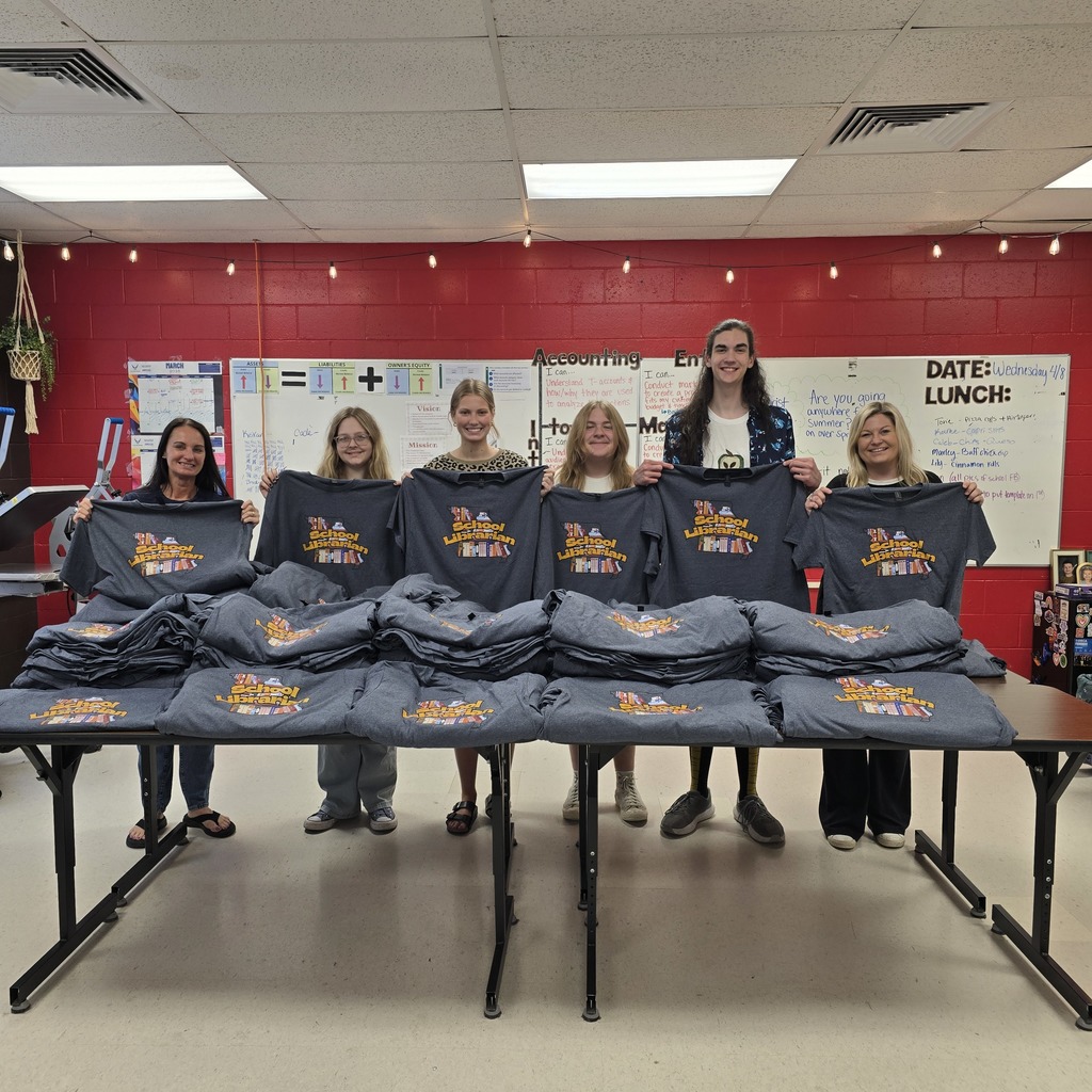 A group of people holds up shirts in front of a table in a classroom.
