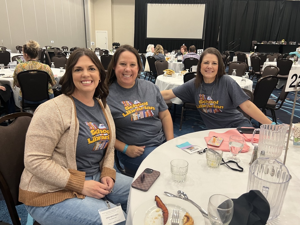 Three women in light blue shirts and light jackets sitting around a table with plates and glasses.