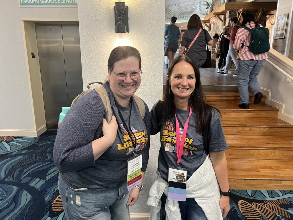 Two women in matching t-shirts and jeans stand smiling for a photo in a building with wooden floors.