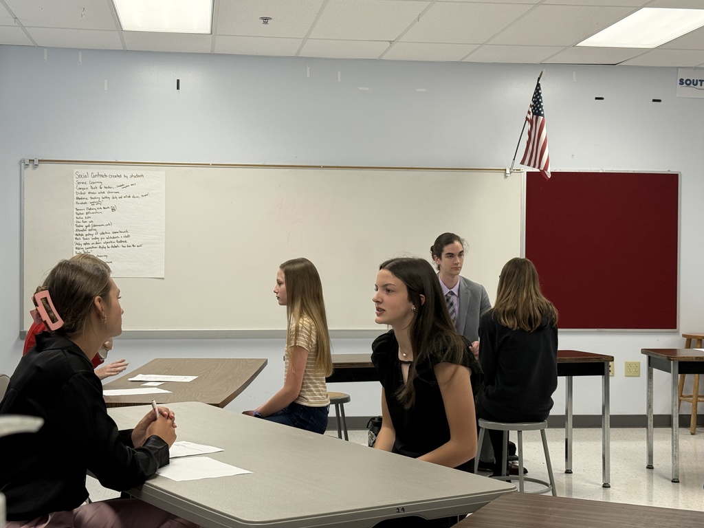 Six people sit at tables in a classroom with a white board and American flag.