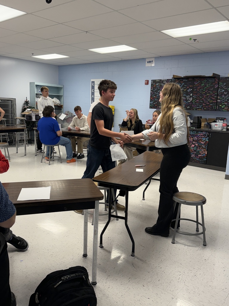 Group of students in a classroom, a student hands a paper to a teacher while standing.