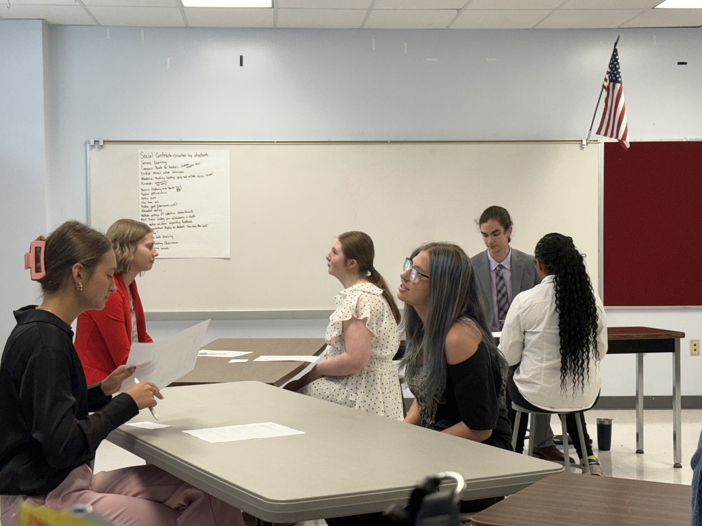 A group of individuals, some seated at tables, engage in a discussion in a classroom. A whiteboard and a flag are visible.