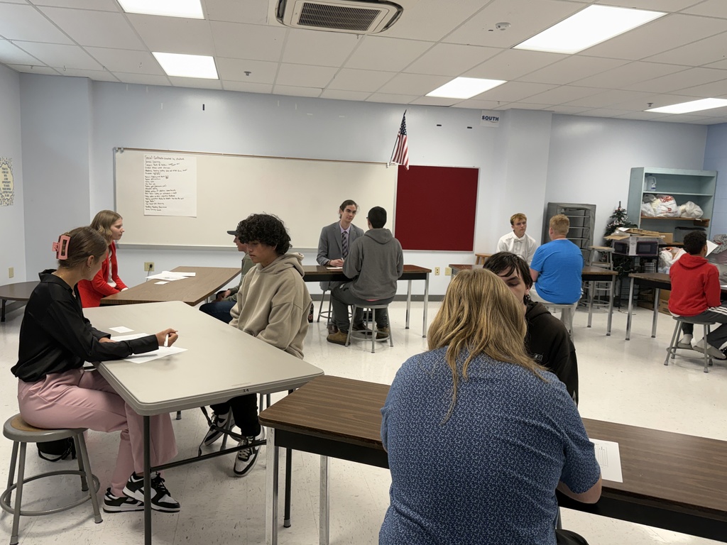 Classroom with students sitting at desks in pairs, one student writing, a whiteboard, and flag in background.